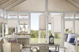 Conservatory dressed in barley-coloured pleated blinds to roof , closed and sides, varying heights, doors open, wood and wicker furniture and striped blue jute rug and cushions.