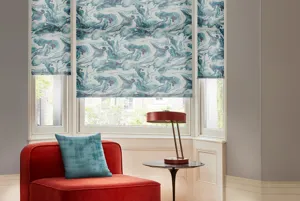 Lounge with narrow but tall bay window dressed in three roller blinds in a white and blue swirling seas pattern.mauve walls, wood floor with black and white rug, low red sofa chair and blue cushion in front of window. 
