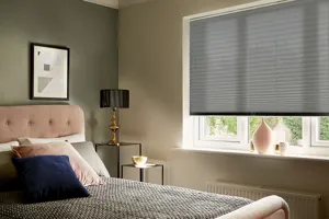 Tight shot of bedroom with one dark and one light brown wall, mid grey pleated blind three quarter lowered at window, bed with dusky pink head and foot, grey bedding, pink, brown and grey accessories.