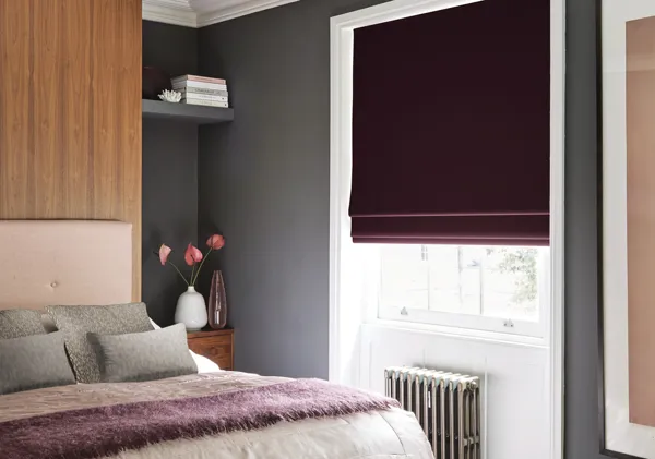 Bedroom with pink and grey bedding, pink headboard against mid-wood wall, grey wall to side with white inset with silver radiator and white sash window dressed with plum chenille Roman blind lowered three quarters.