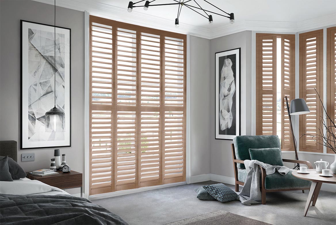 Burley Vintage Rye Shutters have been fitted to tall windows in a bedroom. The Shutters are Natural, Brown coloured while the bedroom is decorated with grey tones.