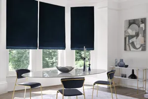 Modern dining room with bay window dressed in three navy blue velvet Roman blinds, oval glass table, metallic gold chairs with blue upholstery, metallic gold side table with blue glassware.