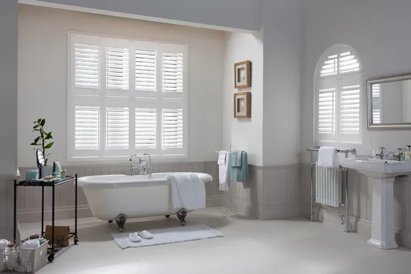 White bathroom with bath under rectangular window dressed in white shutters, and arched window on left wall also in white shutters.