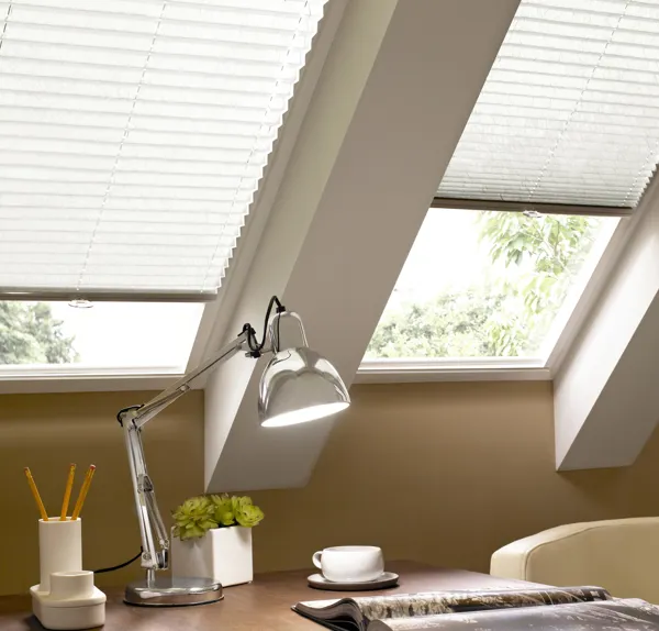 Close up on eaves with desk beneath two skylight windows dressed in off-white pleated blinds one lowered halfway, the other three quarters, silver anglepoise lamp and small plant in white pot, white desk tidy with pencils and a white cup and saucer.