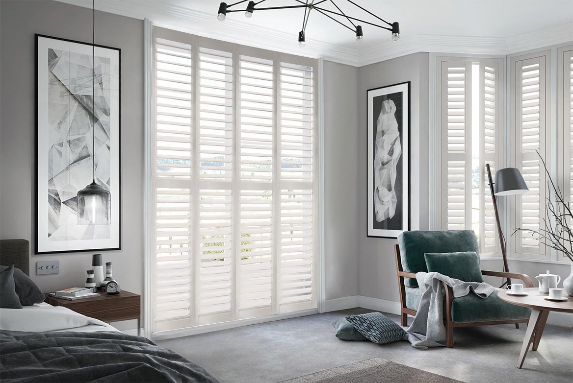 Soft Cream Shutters have been fitted to tall windows in a bedroom. The Shutters are White coloured while the bedroom is decorated with grey tones.