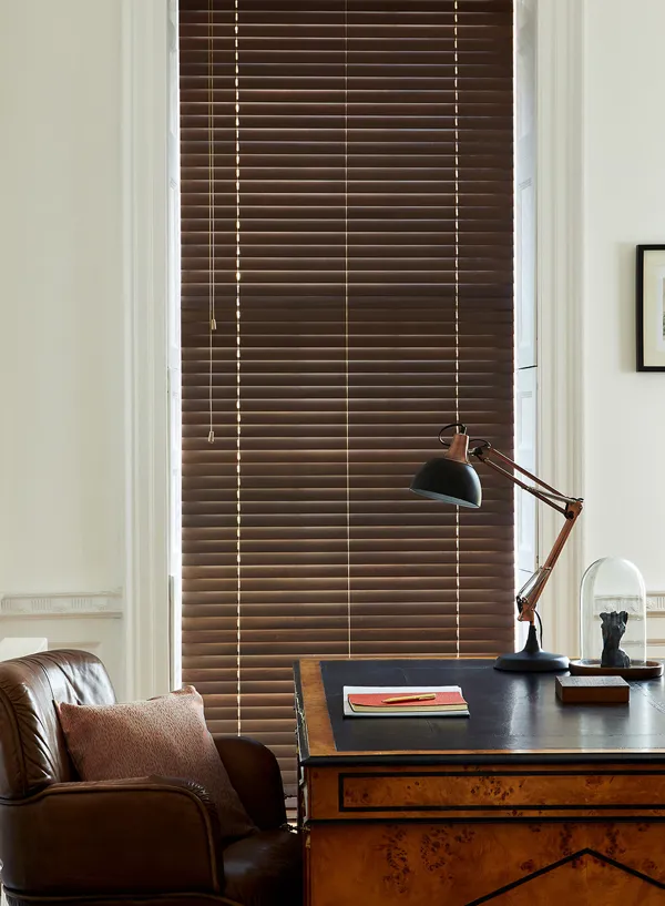 Home office with cream walls, traditional wooden desk, leather tub chair, black anglepoise lamp and tall narrow window dressed in dark wooden venetian blind, lowered, louvres almost totally closed.