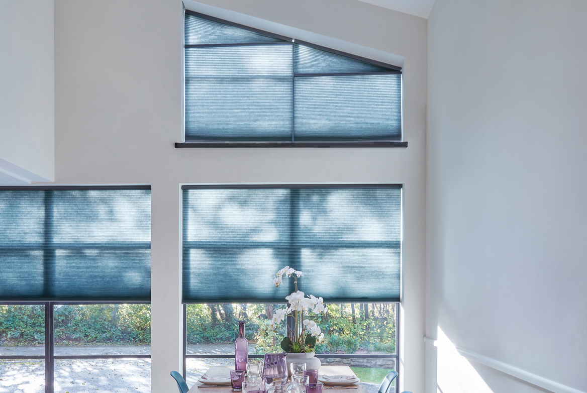 Dining kitchen with double height windows, top window shaped and all dressed in marine blue duette blinds, top closed and others halfway, wooden dining table with blue chairs set for six, white walls filled with light rays.