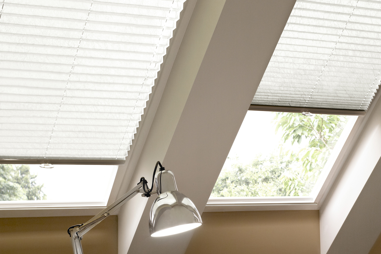 Close up on eaves with desk beneath two skylight windows, silver anglepoise lamp and windows dressed in off-white pleated blinds one lowered halfway, the other three quarters.