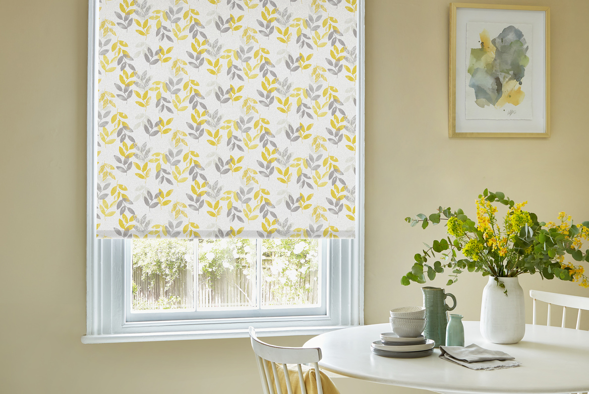 Dining room country chic style with white wood round table and two chairs, white vase with yellow foliage, ochre walls and window dressed in roller blind with grey and yellow leaf pattern
