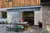 Exterior of split render and wood panel housewith grey striped awning extended over wooden table and benches set for meal, cushions and throws on bench.