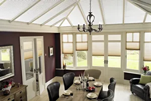 Conservatory with formal dining table, set and side windows dressed in oatmeal pleated blinds at varying  heights and off-white Duette roof blinds.