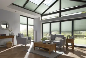 Grey living room with gable end windows and doors, glass roof and patio door to left dressed in pale pine green duette blinds at varying levels, wood floor, grey sofa and chair, wood coffee table and side table all on a grey rug.