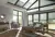 Grey living room with gable end windows and doors, glass roof and patio door to left dressed in pale pine green duette blinds at varying levels, wood floor, grey sofa and chair, wood coffee table and side table all on a grey rug.