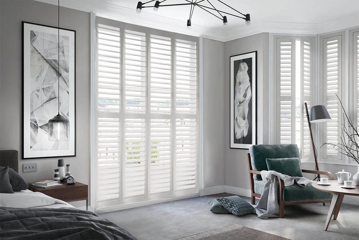 On a series of tall windows Cloudy Skies Shutters have been fitted. The White colour suits the bedroom which is decorated in grey and wooden tones.