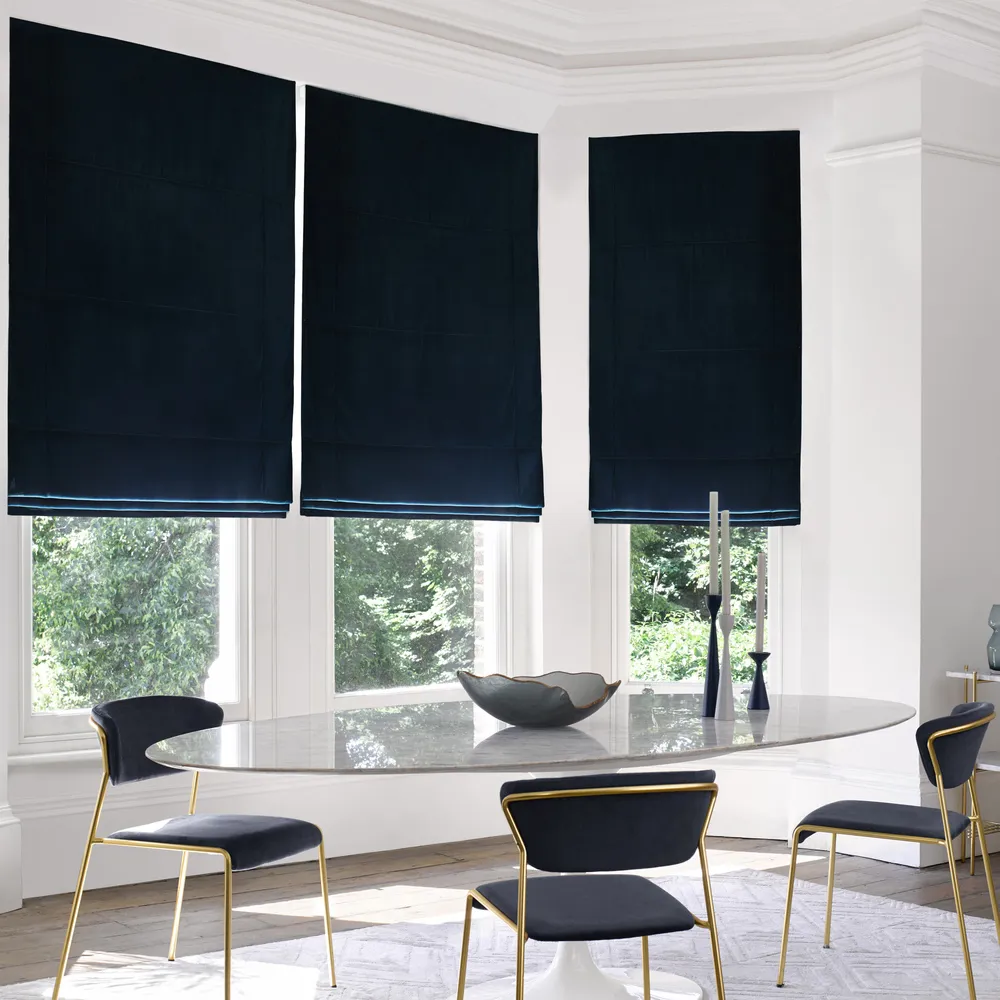 Modern dining room with bay window dressed in three navy blue velvet Roman blinds, oval glass table, metallic gold chairs with blue upholstery, metallic gold side table with blue glassware.