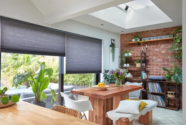 Dining area with door and window combination dressed in blue-grey duette blinds partially lowered, rustic wooden table and benches underneath a roof lantern window.
