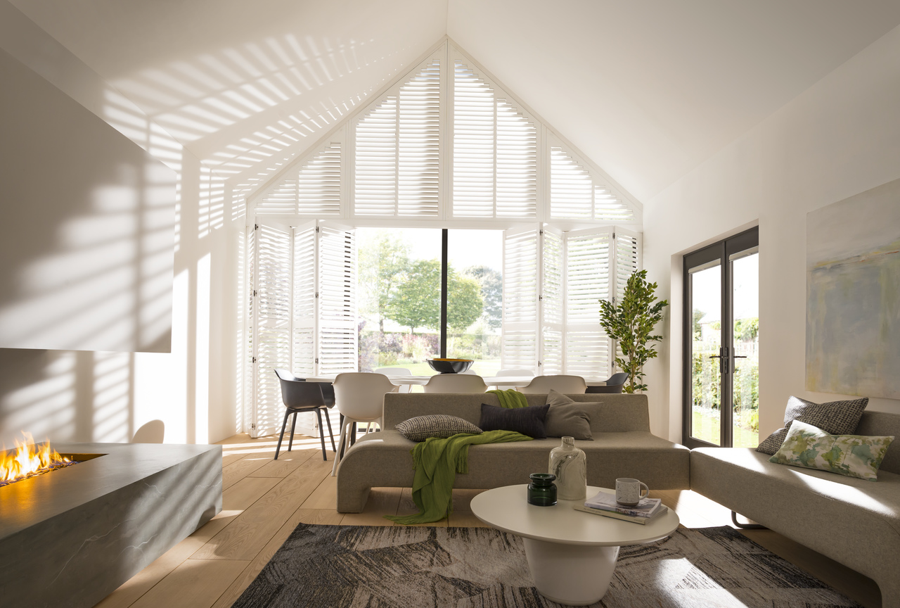 Neutral lounge with feature fireplace, lit, with beige sofas, contemporary table and chairs in front of gable end windows and doors dressed in white shutters.