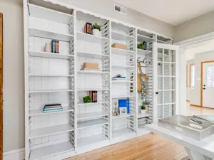 A home office with wooden floor, floor to ceiling white shelving with a mix of empty shelves and books, stationary and plants.