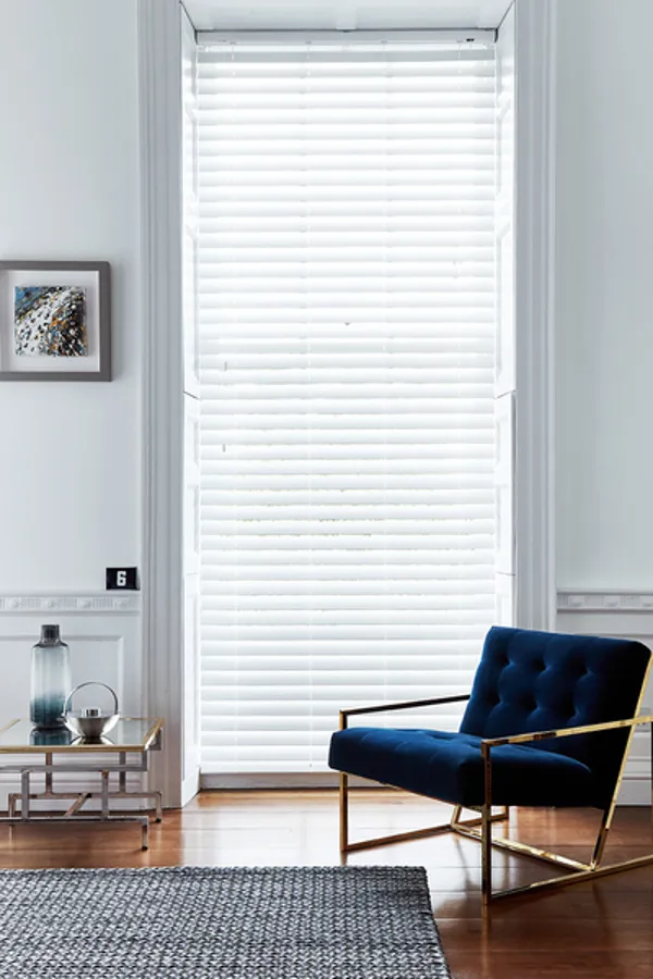 Living room in white with tall narrow window dressed in white wood venetian blind, lowered, slats slightly tilted, polished wood floor, blue marl rug, low blue velvet chair.