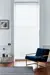 Living room in white with tall narrow window dressed in white wood venetian blind, lowered, slats slightly tilted, polished wood floor, blue marl rug, low blue velvet chair.