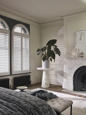 white shutters fitted to an arch shaped window in a bedroom decorated in white with a rug and wooden flooring