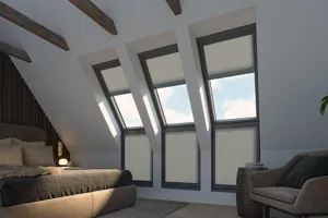 Bedroom in white with sloped ceiling and wall with three skylight windows above three standard windows all dressed in cream duette blinds, skylight blinds raised, lower window linds extended, brown bedding, brown chair in corner.