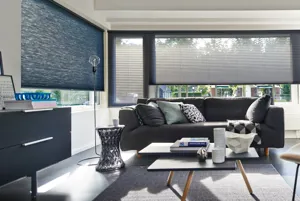 Living room with white walls, two wide windows and one narrow on adjacent walls, all dressed in smoky blue duette blinds, lowered three quarters, grey sofa, two-tiered coffee table with grey surface, on a grey herringbone-weave rug, grey floor, dark grey sideboard, grey drum-style side table.