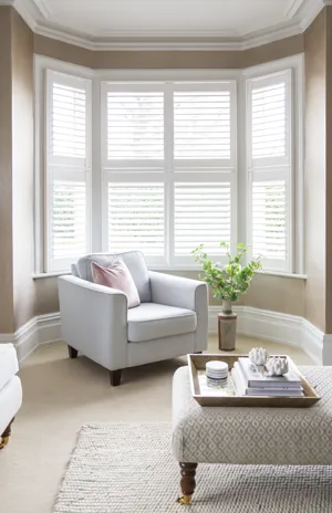 Mid beige living room with cream carpet, cream rug under a cream and wood footstool, cream upholstered chair in the arc of a bay window, which is dressed in four white tier on tier shutters, closed, with louvres open.