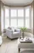 Mid beige living room with cream carpet, cream rug under a cream and wood footstool, cream upholstered chair in the arc of a bay window, which is dressed in four white tier on tier shutters, closed, with louvres open.