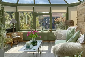 Orangery with dark olive frame, pale wood floor, sofa and coffee table with pink flowers and pale beige roller blinds, raised.
