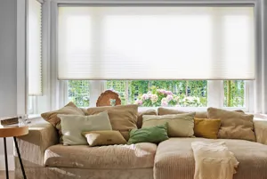 Living room with white walls, deep bay window with wide central window, all dressed in beige duette blinds, lowered three quarter, dark beige cord L-shaped sofa in bay recess with cushions in shades of brown, cream and green, mid beige throw on seat.